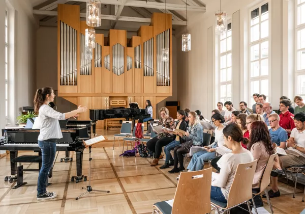 Mach Kirchenmusik R Ein Kirchenchor sitzt im Halbkreis vor einer Dirigentin, im Hintergrund ist eine große Orgel